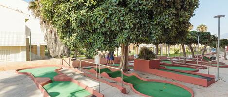 Un campo de mini golf con varios hoyos, rodeado de una zona pavimentada y un árbol grande.