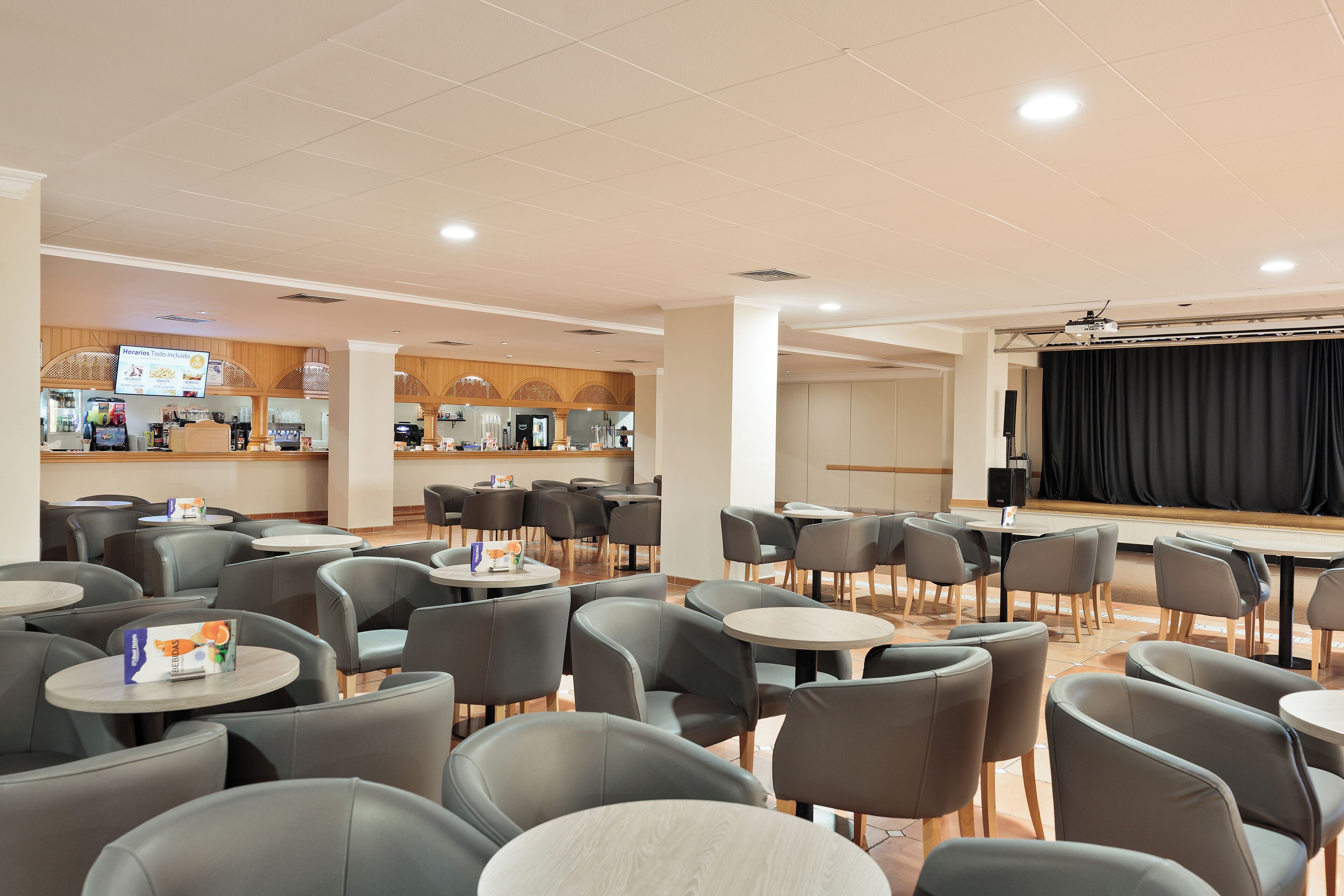 A spacious indoor area with multiple seating arrangements, including round tables and chairs, and a stage with a black backdrop.