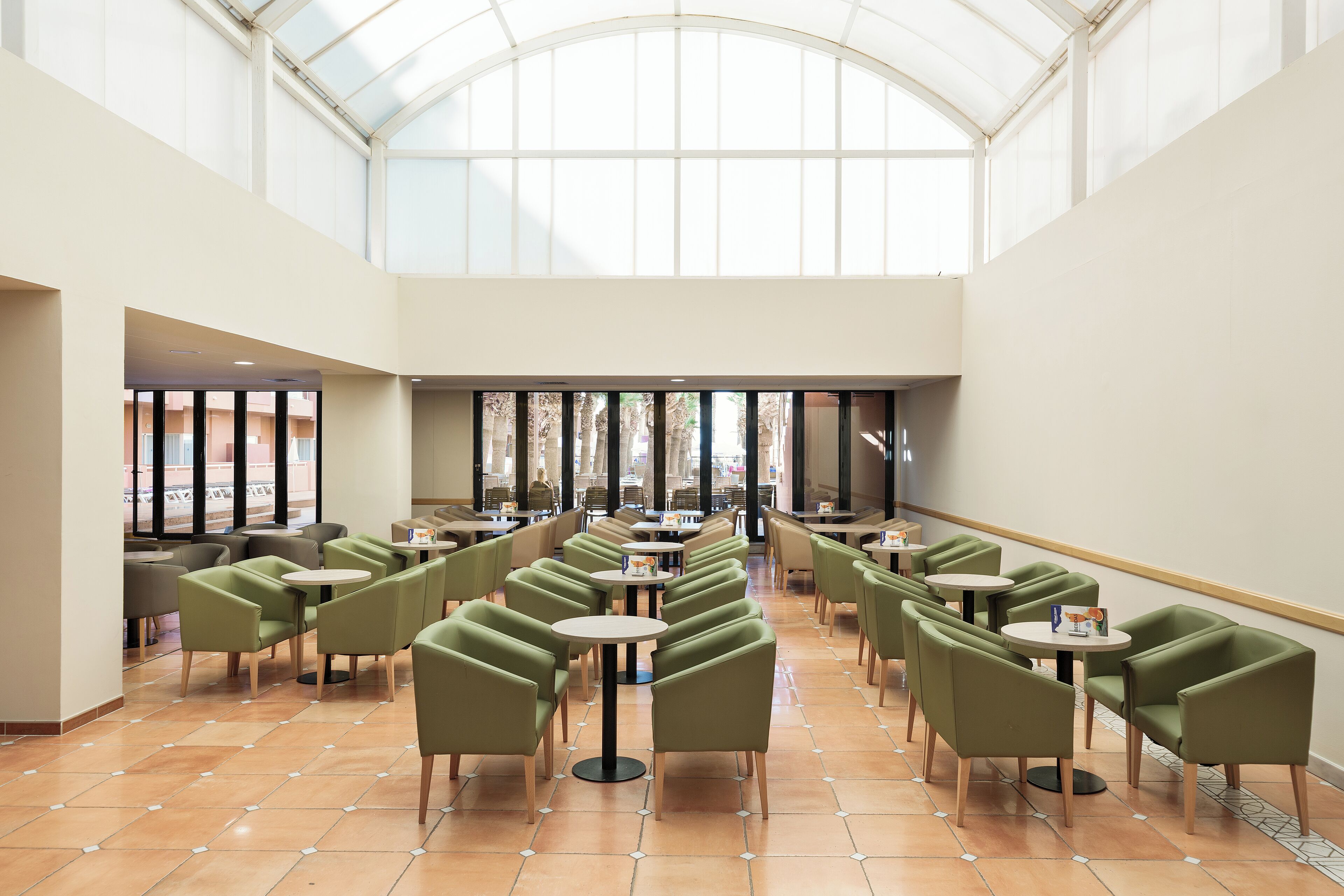 A spacious indoor area with multiple tables and chairs arranged in rows, large windows allowing natural light, and a high ceiling with a skylight.
