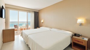 A hotel room with two beds, a desk, a chair, a nightstand with a phone, and a view of the sea through large windows.