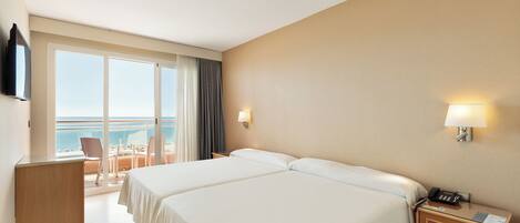 A hotel room with two beds, a desk, a chair, a nightstand with a phone, and a view of the sea through large windows.