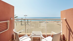 A balcony with white chairs and a table, overlooking a beach with umbrellas and people.