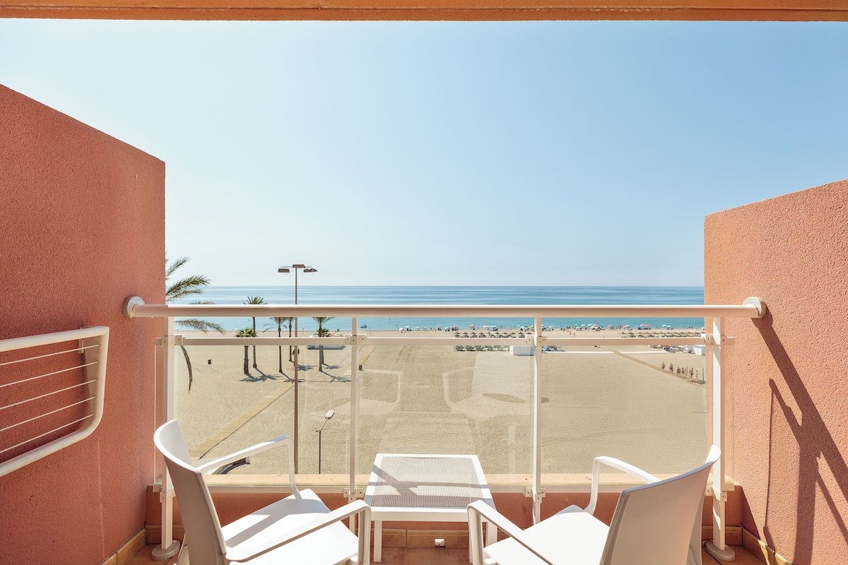 Un balcón con vistas a la playa, sillas blancas y una mesa.