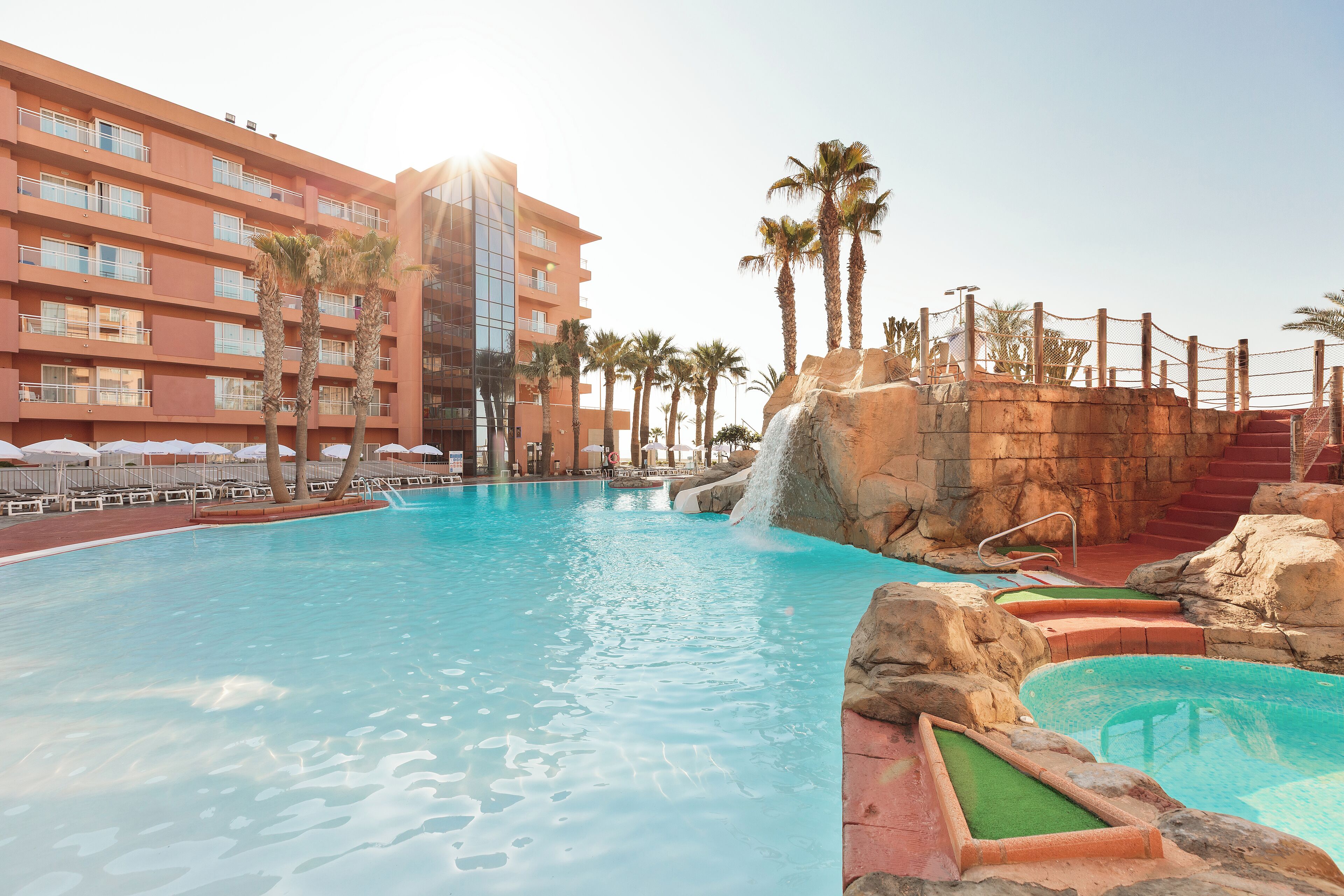 A swimming pool with a waterfall feature, surrounded by palm trees and a hotel building.