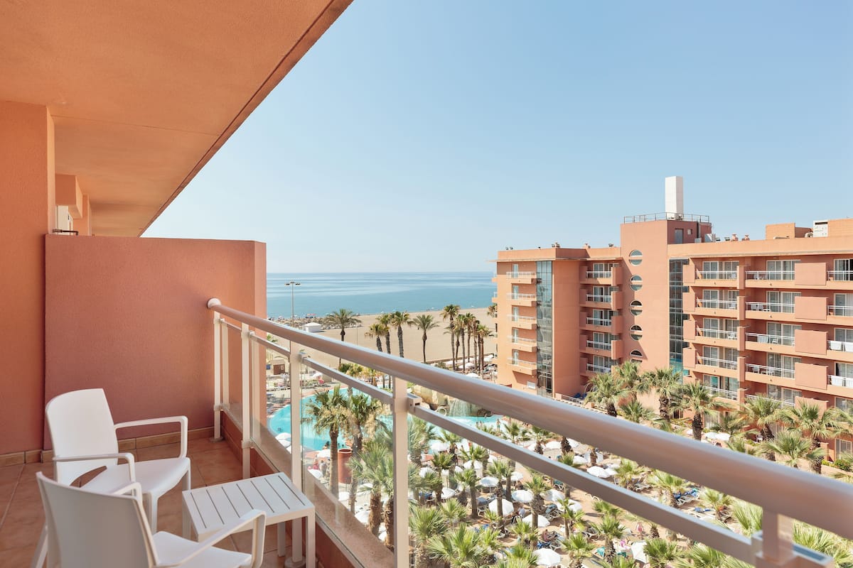 Un balcón con sillas y mesa blancos, con vistas a una zona de piscina con palmeras y una playa.