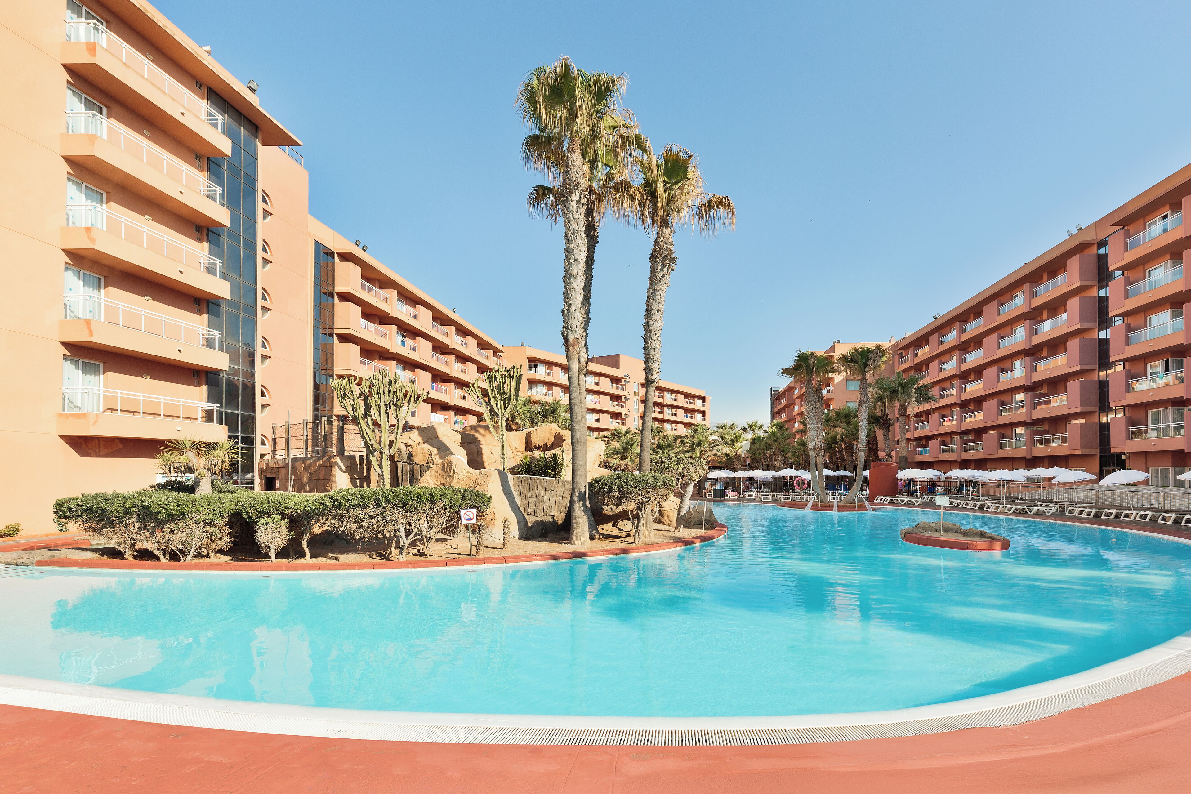 Un hôtel doté d’une piscine entourée de palmiers, avec un ciel bleu et dégagé.