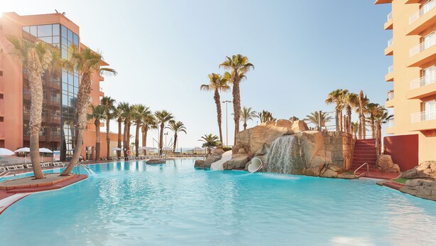 A swimming pool with palm trees, a waterfall, and modern buildings.