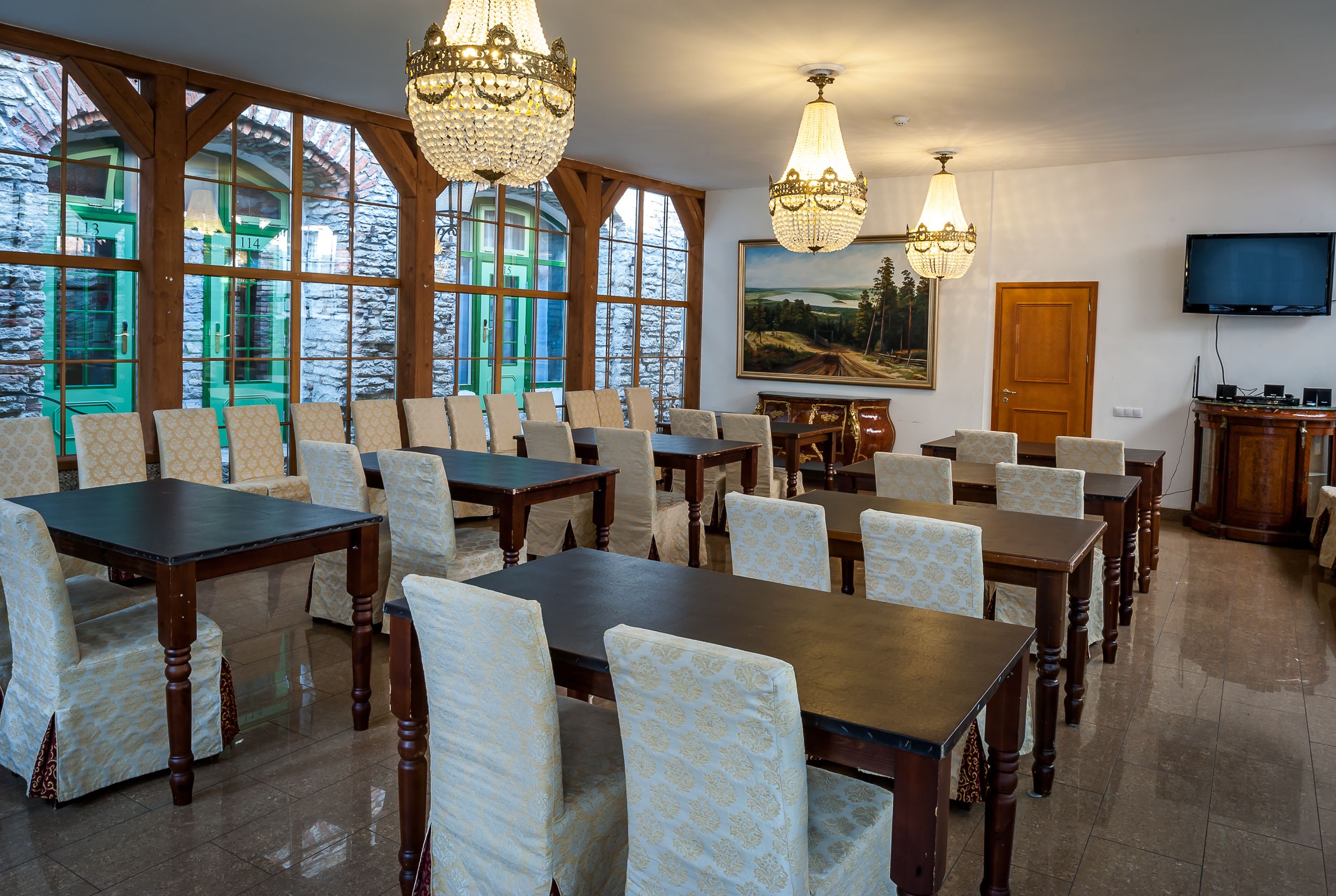 A dining area with multiple tables and chairs, large windows with a view of trees, and chandeliers.