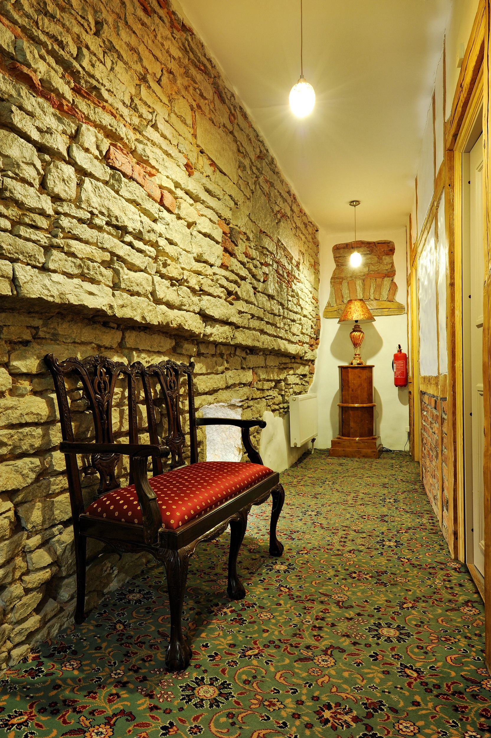 A narrow hallway with a stone wall, a wooden bench with a red cushion, a patterned carpet, and a lamp.