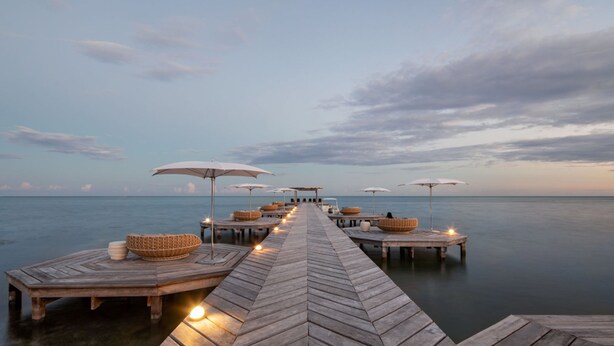 Un muelle de madera que conduce a una serie de plataformas flotantes con sombrillas y zonas de estar, con vista a aguas tranquilas.
