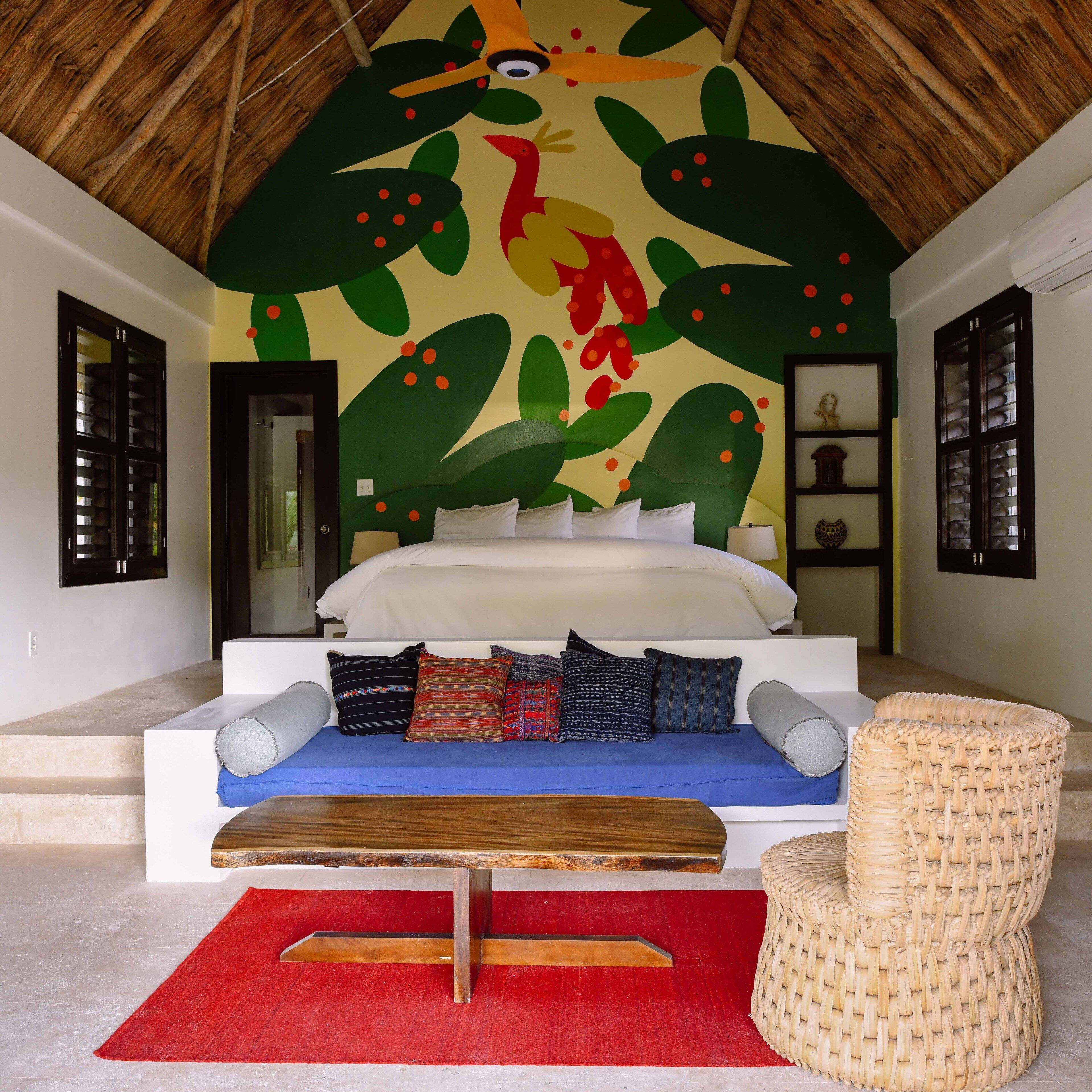 A bedroom with a bed, pillows, a red rug, a woven chair, and a wall mural.