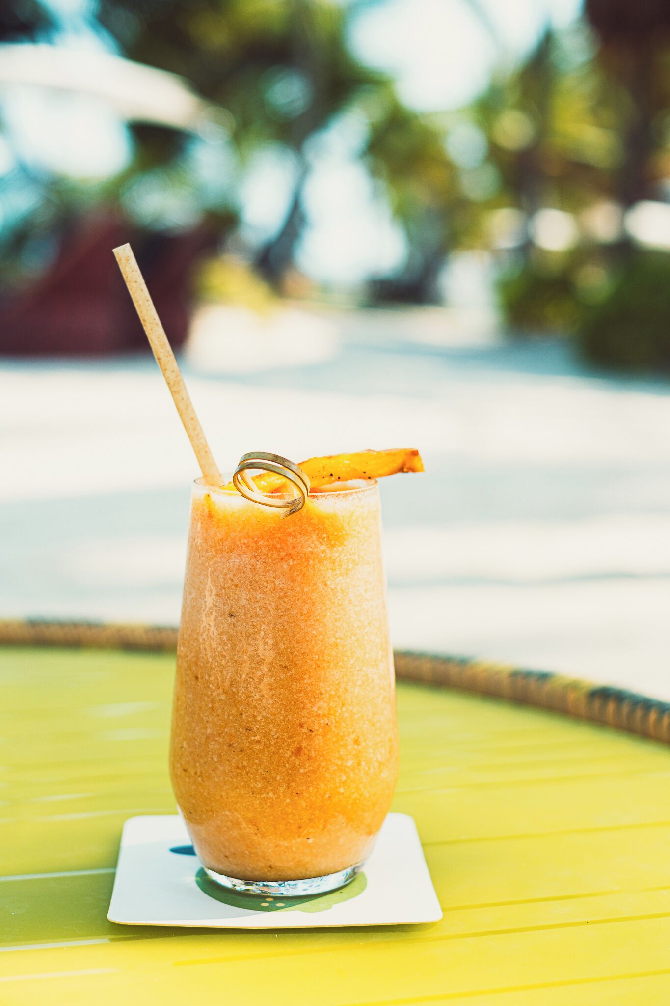 A tall glass of orange smoothie with a straw and a slice of orange on a white coaster, placed on a yellow table.