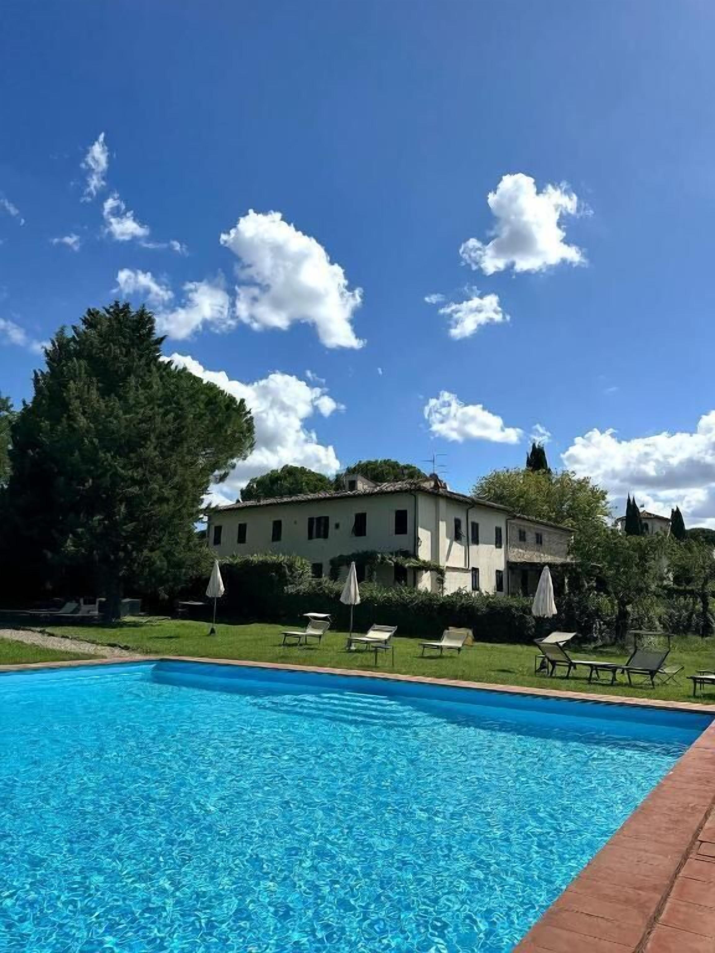 A swimming pool with lounge chairs and umbrellas, a large building in the background, and a clear blue sky.