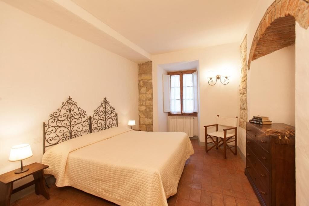 Un dormitorio con una cama, un aparador de madera, una silla, una mesa y un ventanal con cortinas.