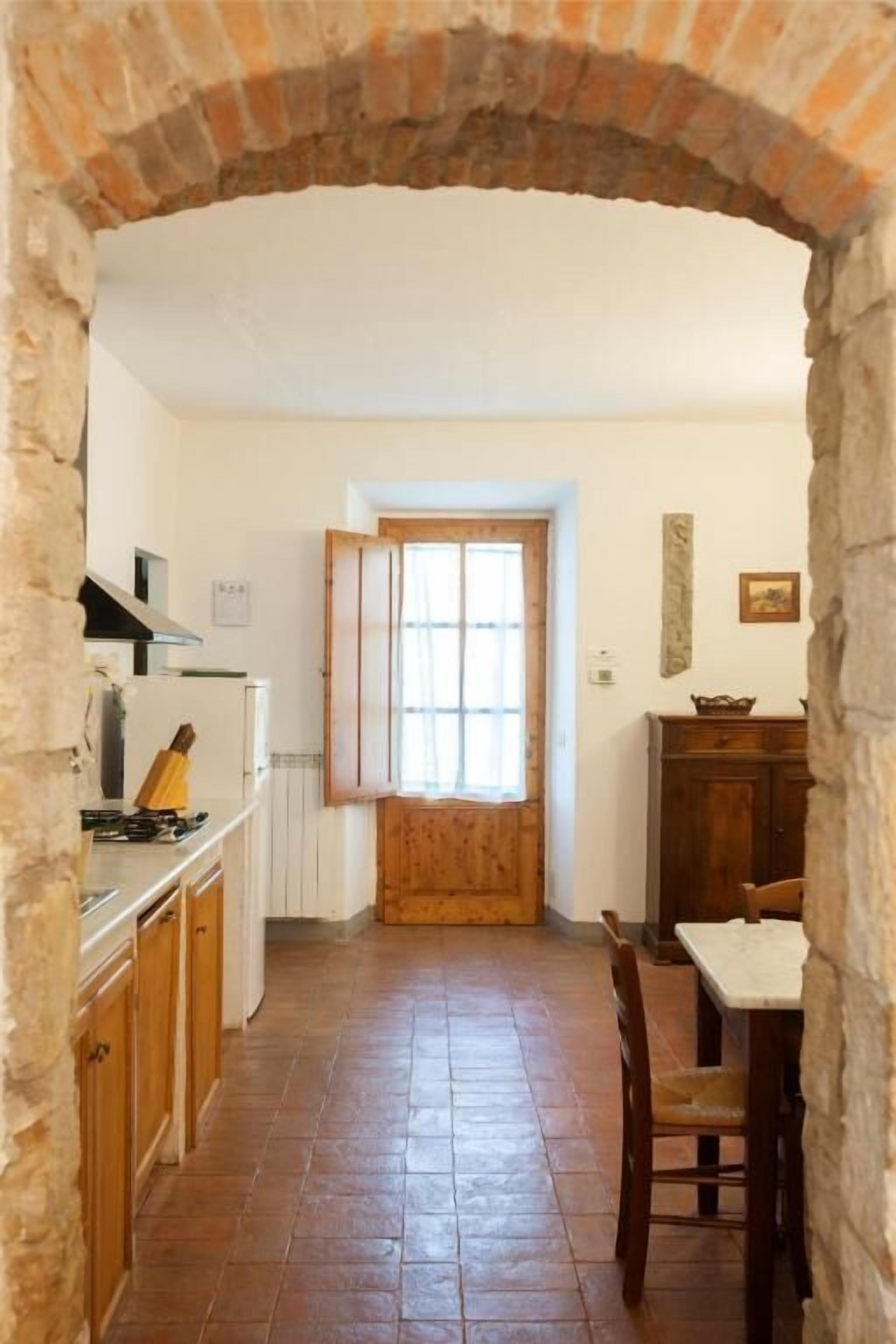 A narrow kitchen with a brick archway, wooden cabinets, a white refrigerator, and a window with wooden shutters.