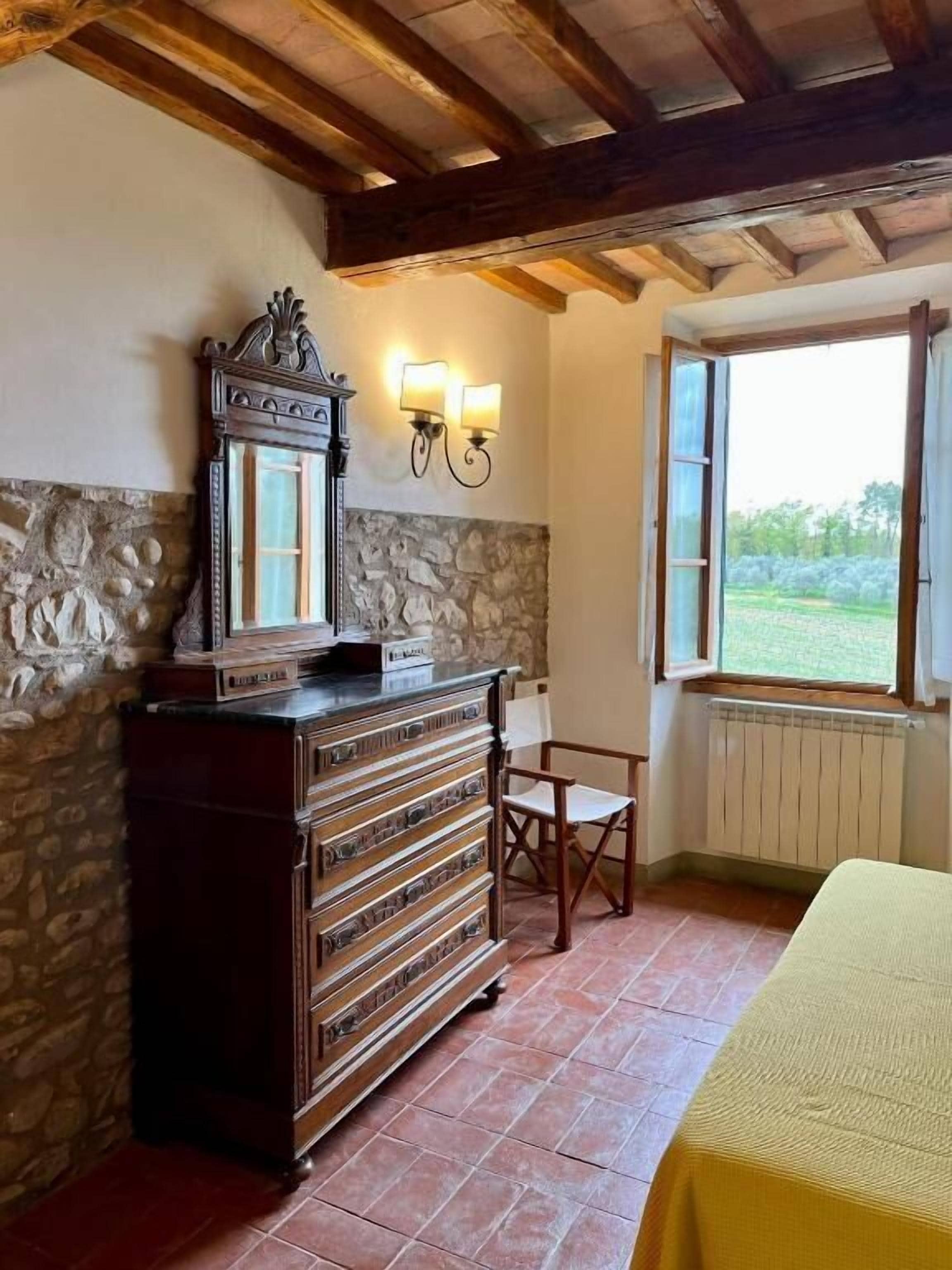 Quarto com parede de pedra, cómoda de madeira, duas luminárias fixadas na parede, uma cadeira, uma mesa com toalha amarela e uma janela com persiana aberta.