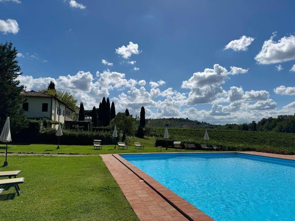 Une piscine, avec un ciel bleu clair et quelques nuages, entourée d’une pelouse où sont disposés des transats et des parasols.