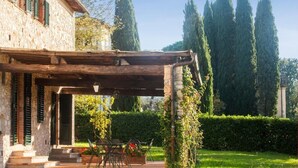 A stone building with a covered patio, outdoor seating, and tall trees in the background.