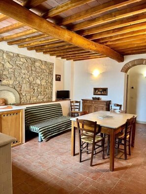 Ein rustikales Interieur mit freiliegenden Holzbalken, einer Steinwand, einem Fliesenboden, einem Esstisch mit Stühlen und einem gestreiften Sofa.