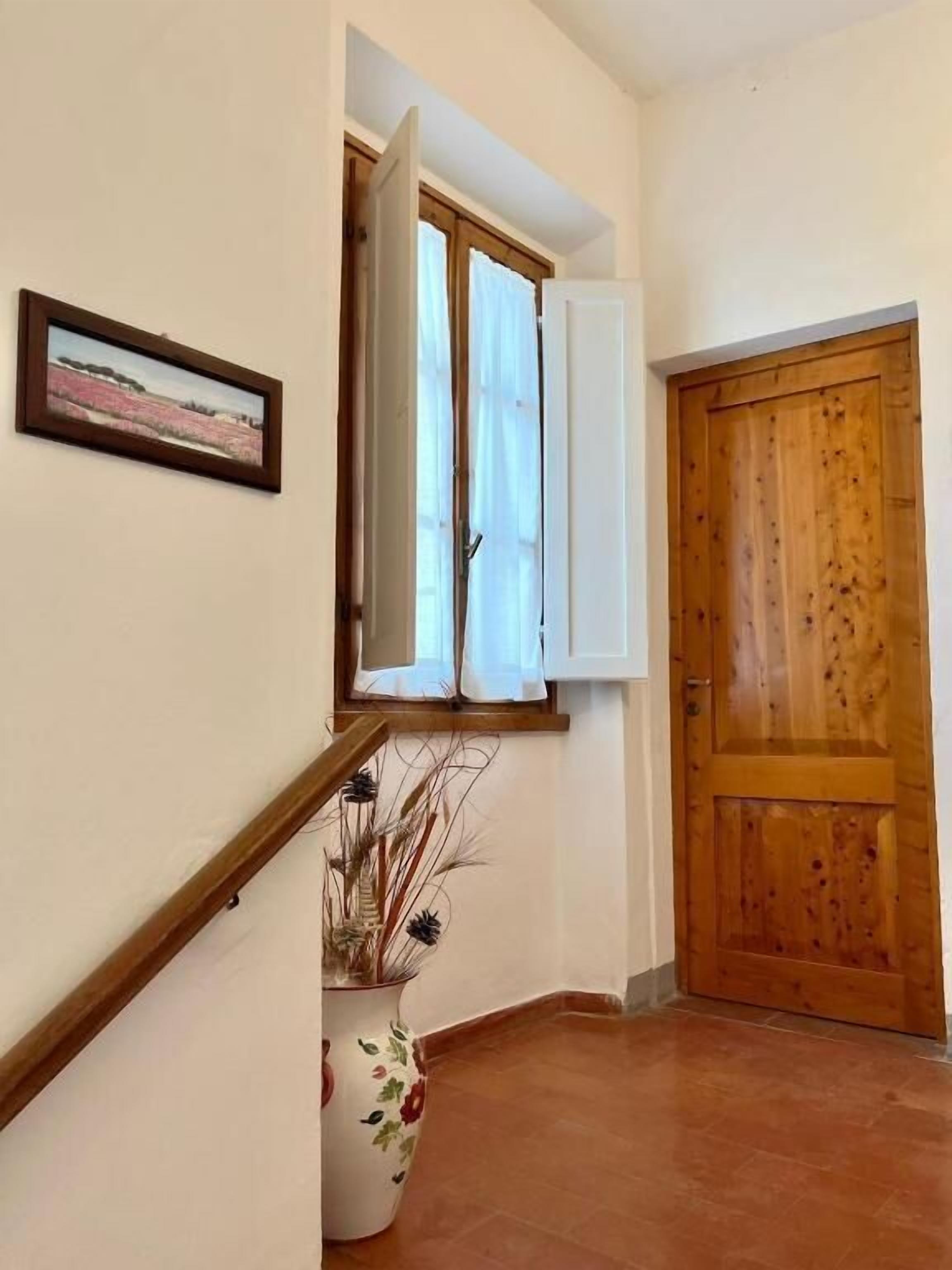 Ein Korridor mit einer Holztür, einem Fenster mit weißen Vorhängen, einem gerahmten Bild an der Wand und einer Topfpflanze.