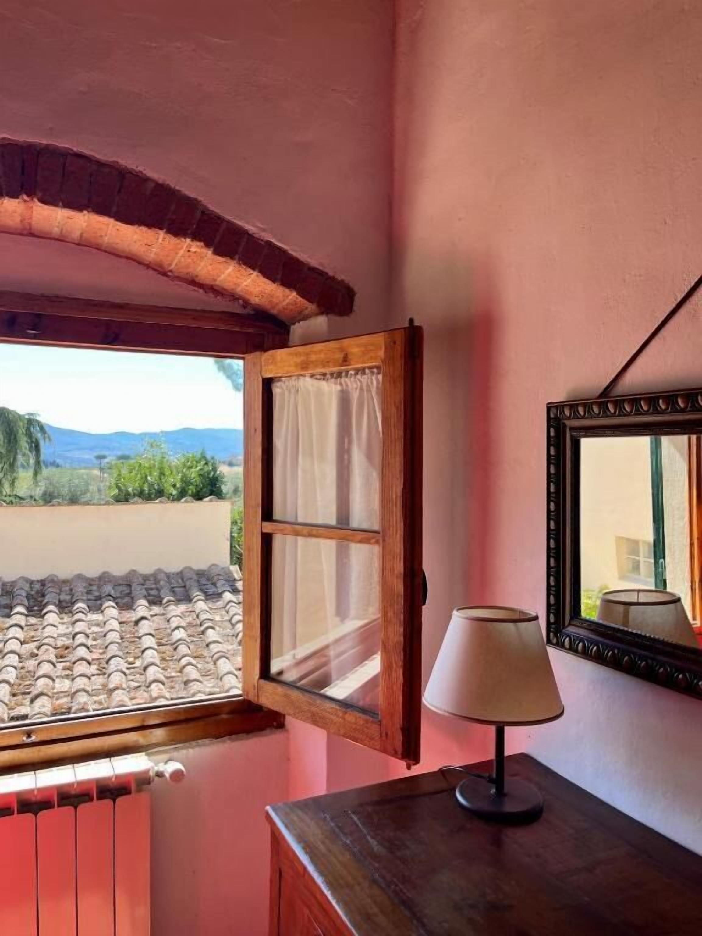 A room with a wooden-framed window, a table with a lamp, and a mirror on the wall.