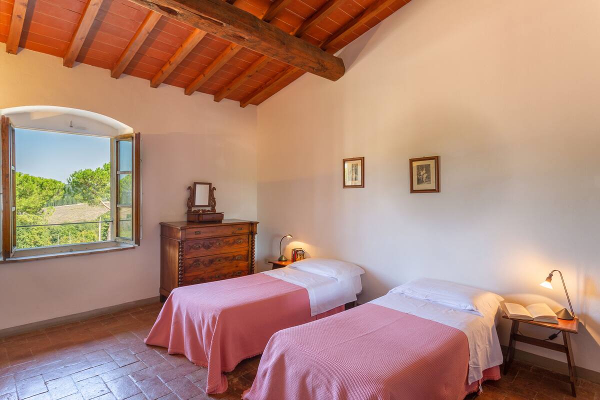 A bedroom with two beds, a wooden dresser, a nightstand with a lamp, and a window with a view of greenery.