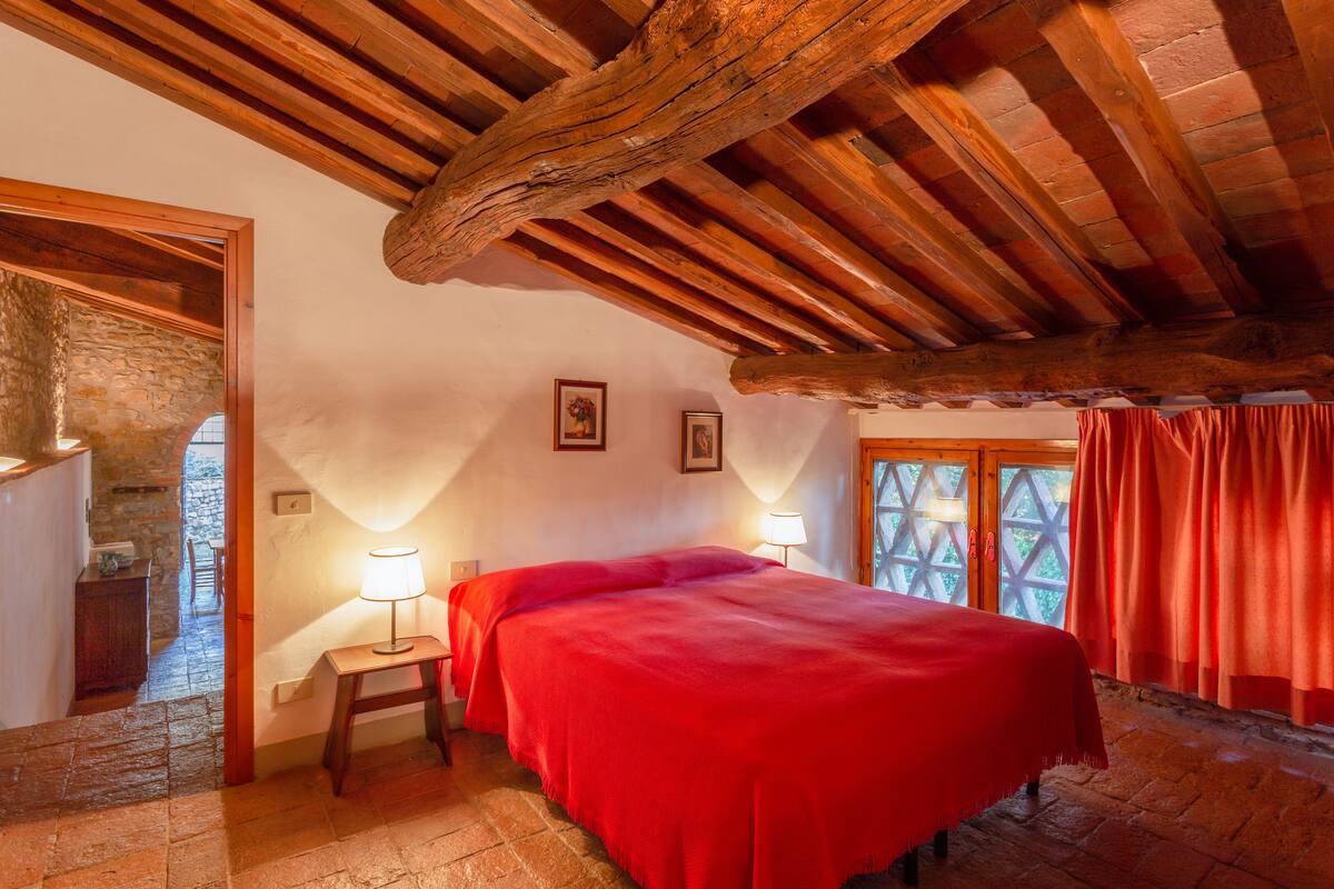 A cozy room with a bed, wooden beams, stone walls, and a window with curtains.