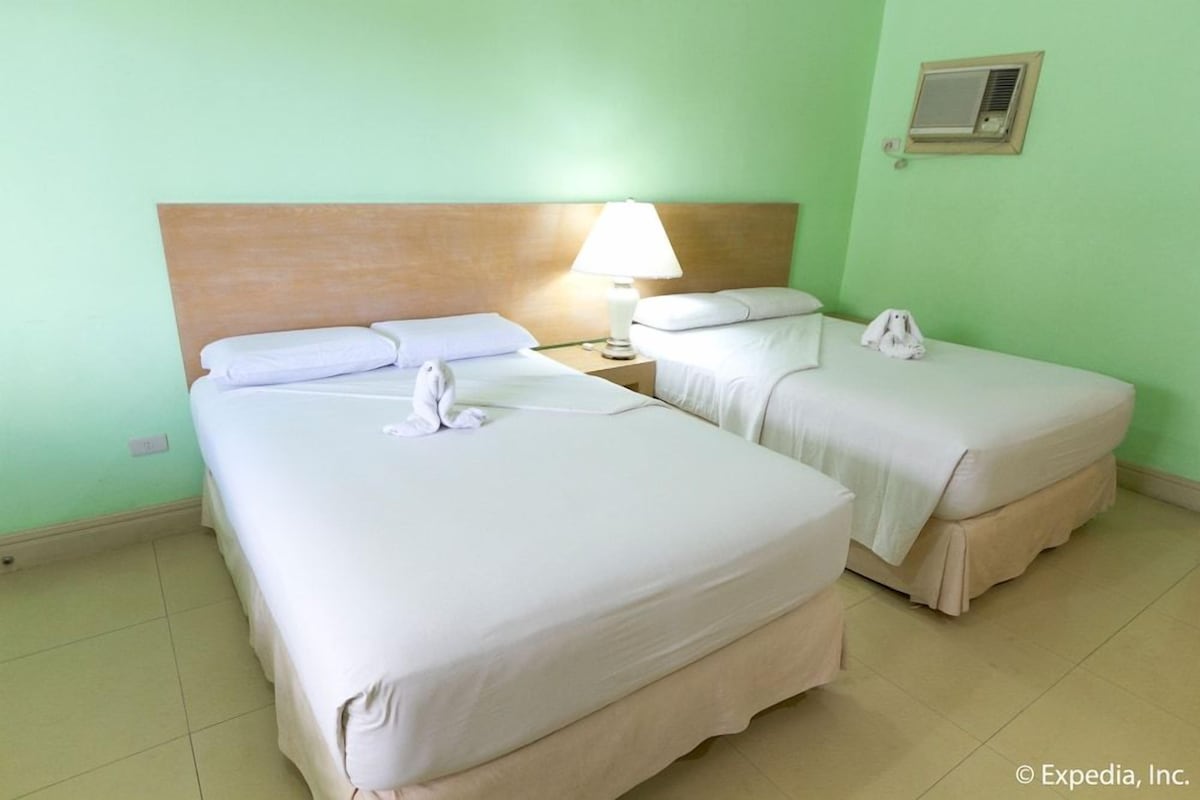A hotel room with two beds, a nightstand with a lamp, and a wall-mounted air conditioner.