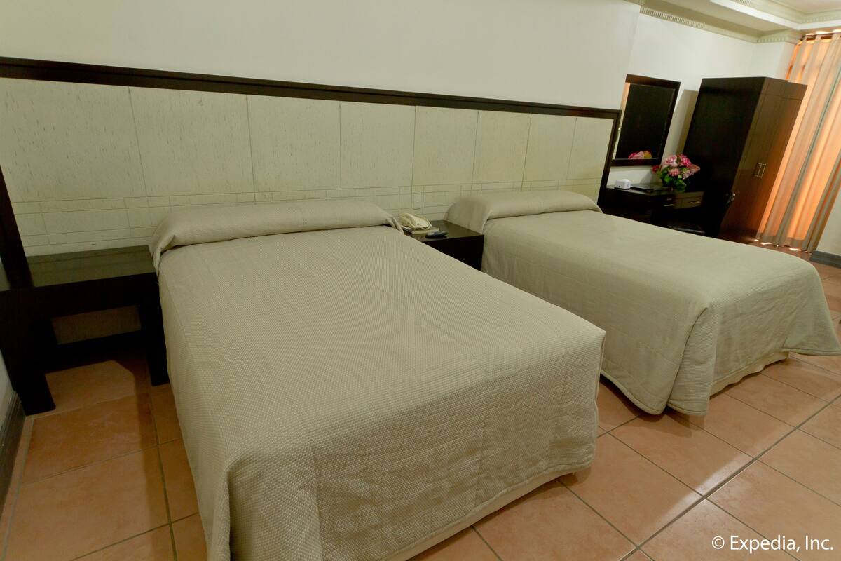 Quarto de hotel com duas camas, uma mesa de cabeceira com telefone, um espelho e um armário com um vaso de flores.