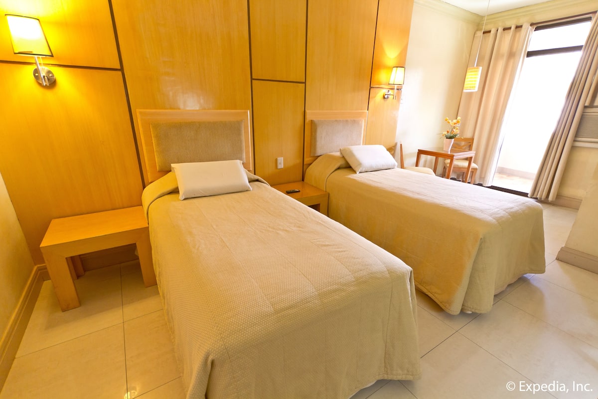 Quarto de hotel com duas camas de solteiro, uma mesa de cabeceira de madeira, uma pequena mesa com uma cadeira e uma janela com cortinas.