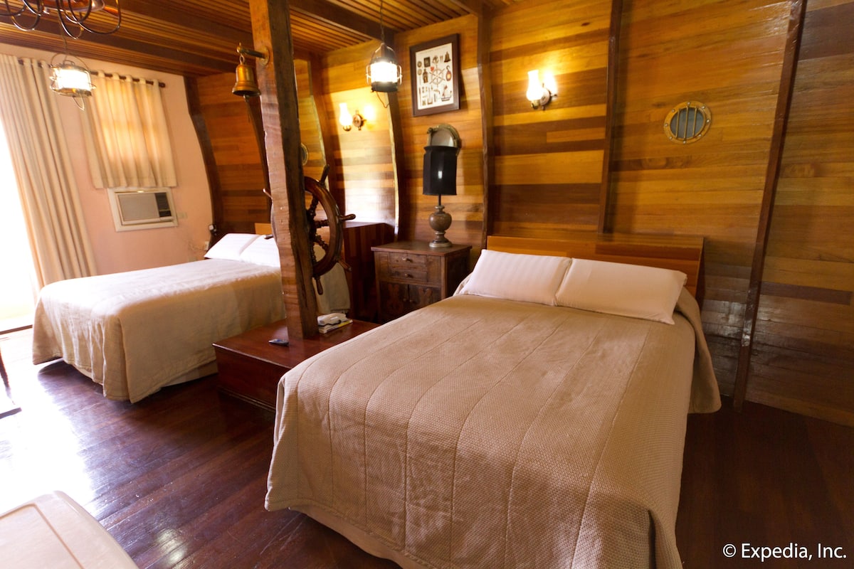 Quarto de madeira com duas camas, uma mesinha de cabeceira, um abajur e um relógio de parede.