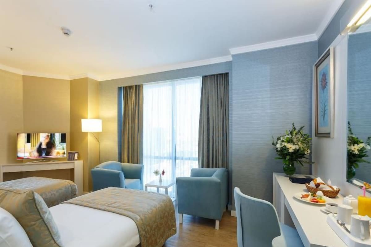 A hotel room with a bed, two chairs, a desk with breakfast items, a TV, and a window with curtains.