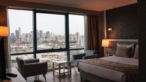 A hotel room with a large window offering a city view, a bed, two armchairs, a small table, and a nightstand with a lamp. - Byotell Istanbul (Istanbul)
