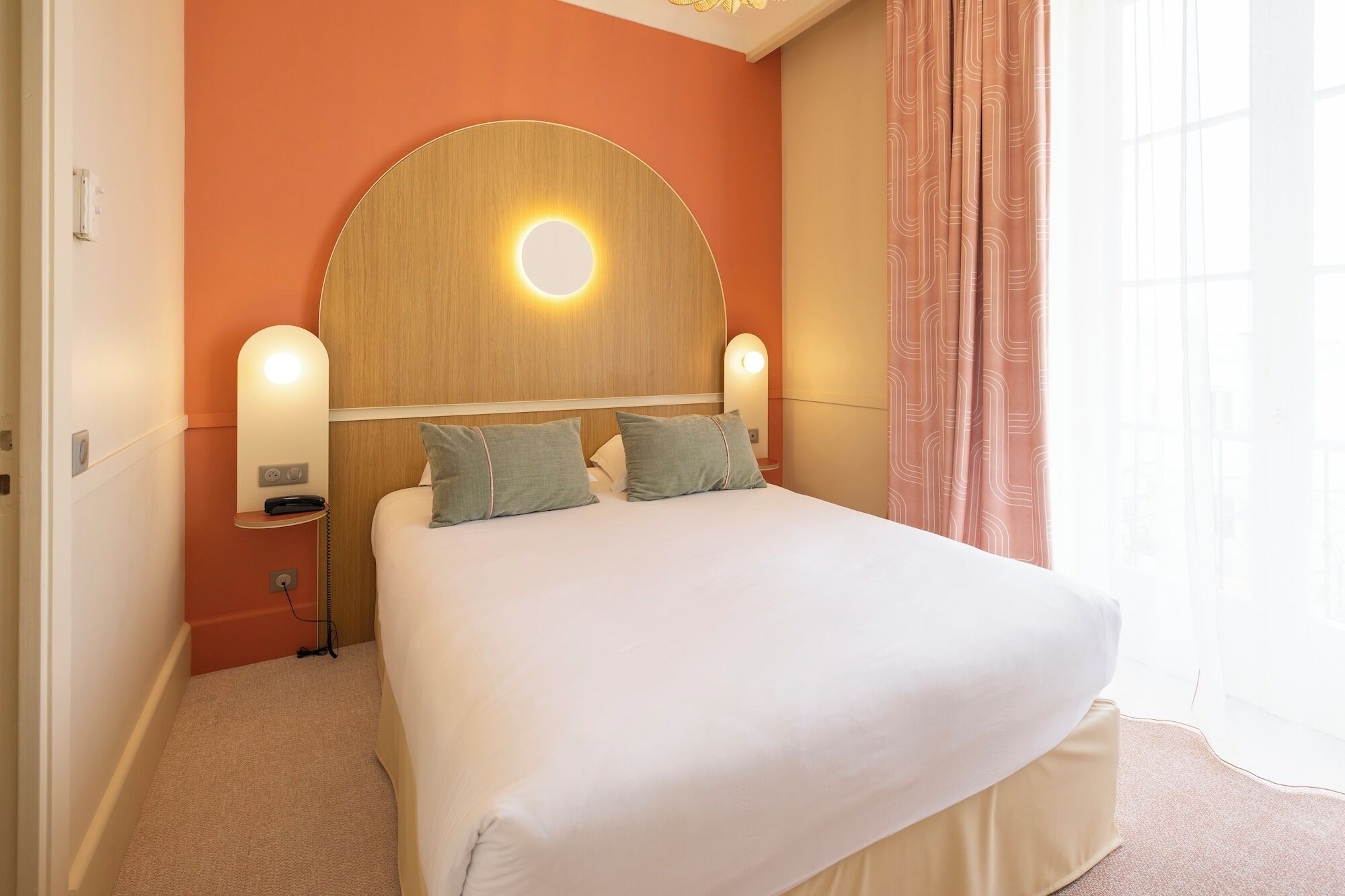 Habitación de hotel con una cama grande, dos lámparas de noche, un espejo redondo sobre la cama y una ventana con cortinas.