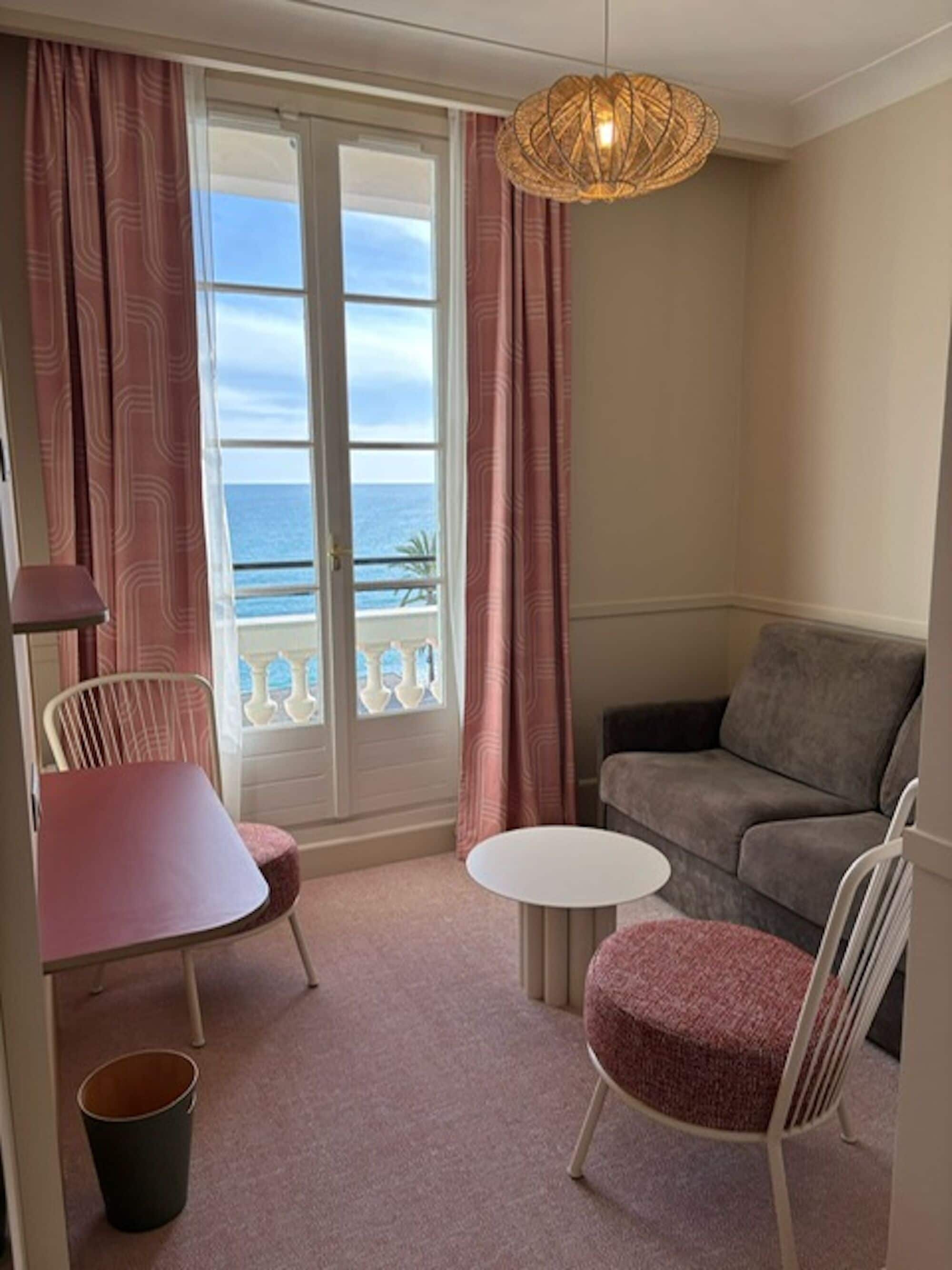 A room with a sofa, two chairs, a small table, a trash bin, and a view of the ocean through a window with curtains.