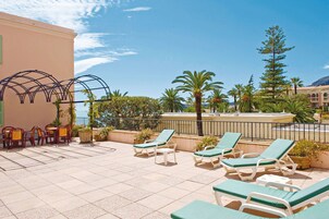 Terraza en la azotea con sillones y mesas, rodeada de plantas en macetas y palmeras, con vistas a un cuerpo de agua y edificios.