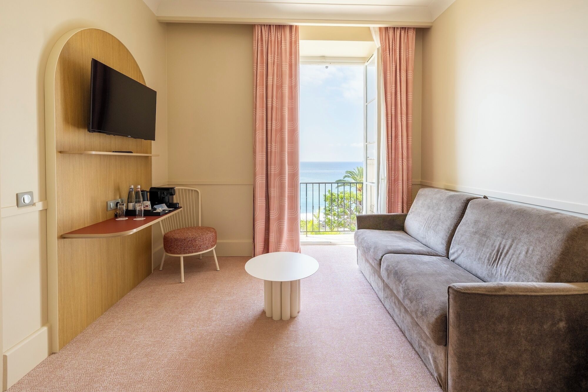 A hotel room with a sofa, a small table, a chair, a flat-screen TV mounted on a wooden panel, a balcony with curtains, and a view of the ocean.