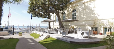 An outdoor patio area with tables and chairs, a large tree, and a view of the sea.
