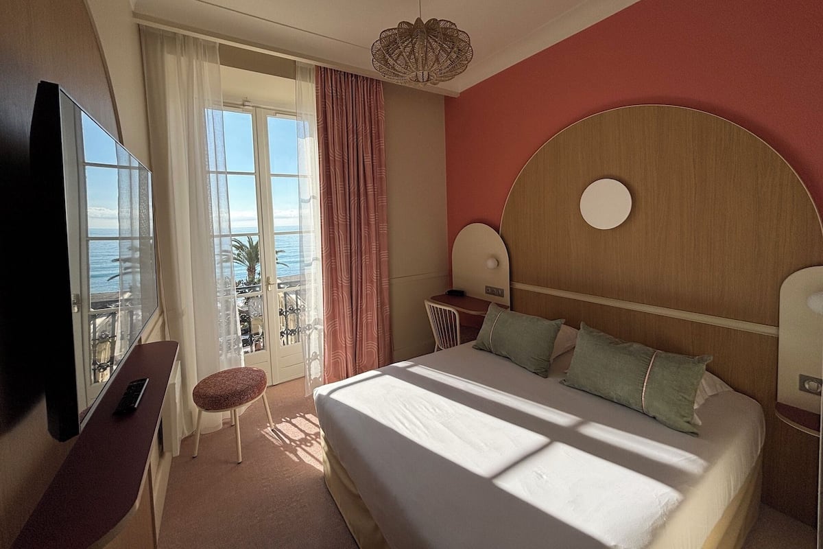 Camera d'albergo con un letto, una scrivania con una sedia, una televisione e un balcone con tende, con vista sul mare.