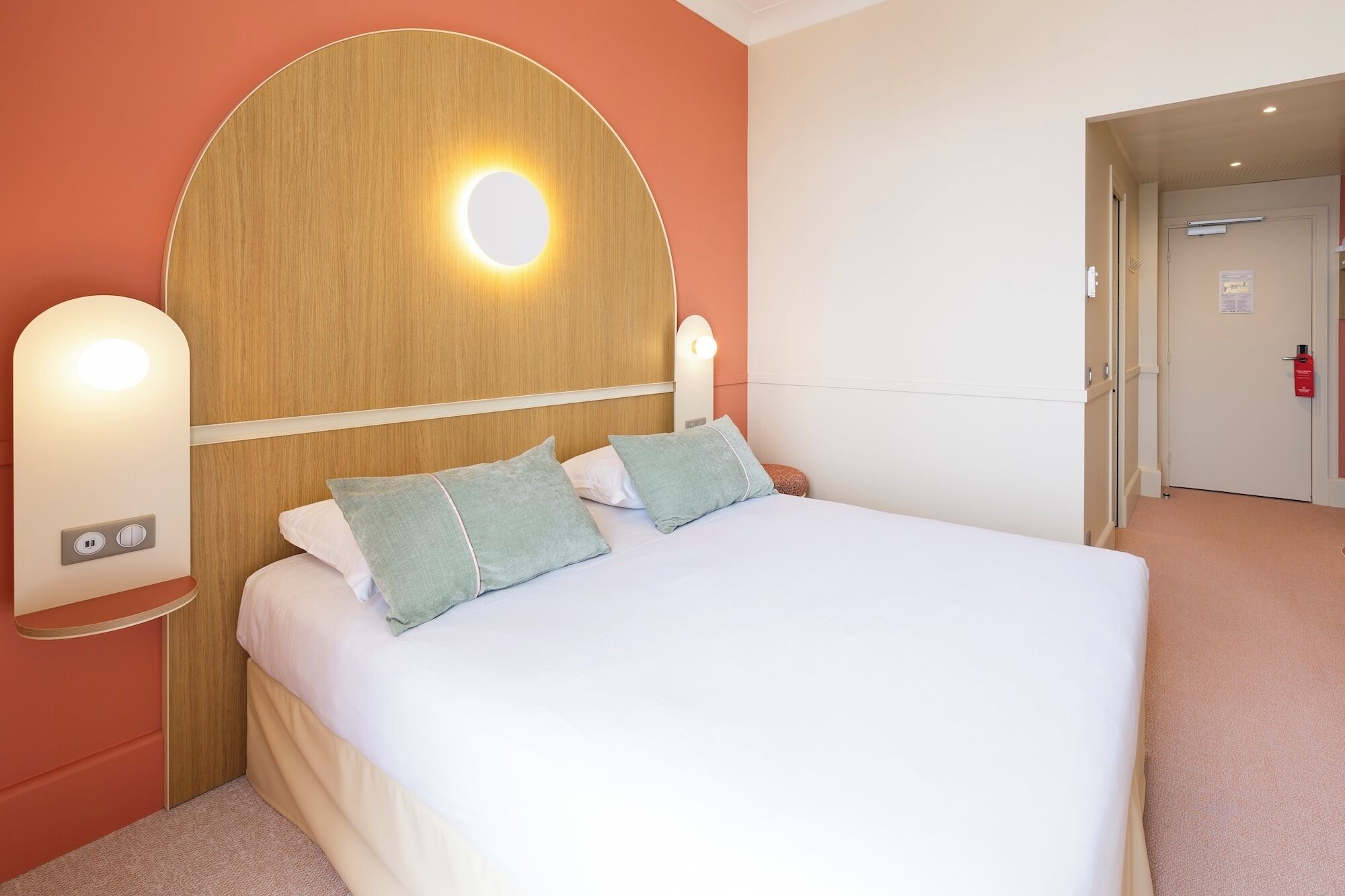 Ein Hotelzimmer mit einem Bett, zwei Wandleuchten, einem Holz-Kopfteil und einer Tür rechts.