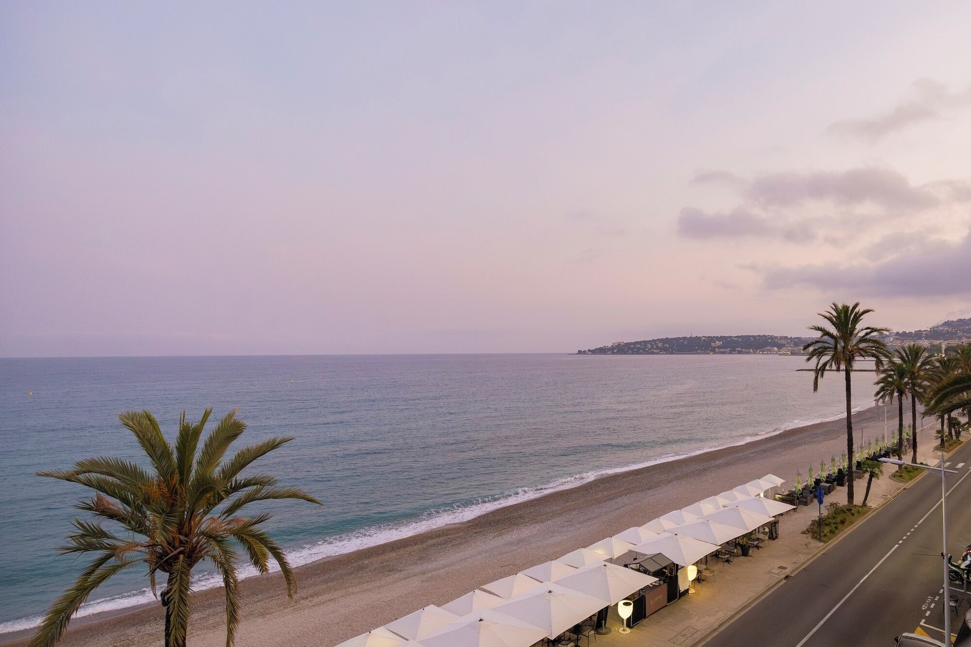 A beachside view with a road lined with umbrellas and palm trees.