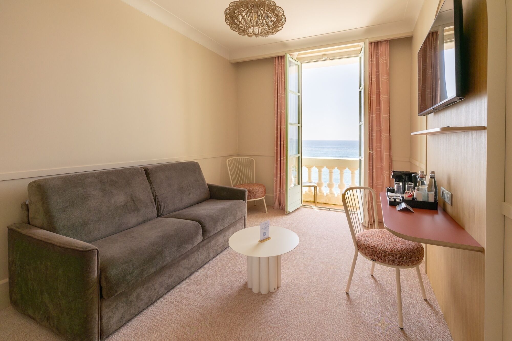 Une chambre d’hôtel avec un canapé, une petite table ronde, une chaise, une télévision et un balcon avec des rideaux.