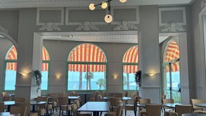 A spacious dining area with multiple tables and chairs, large arched windows with striped awnings, and a ceiling with a modern light fixture.