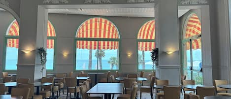 Un comedor espacioso con varias mesas y sillas, grandes ventanas de arco con toldos a rayas y un techo con una lámpara moderna.