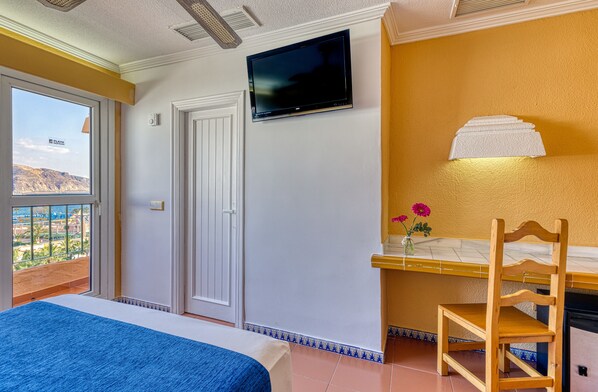 Een hotelkamer met een bed, een aan de muur bevestigde televisie, een houten stoel, een tafeltje met bloemen en een balkon met uitzicht.