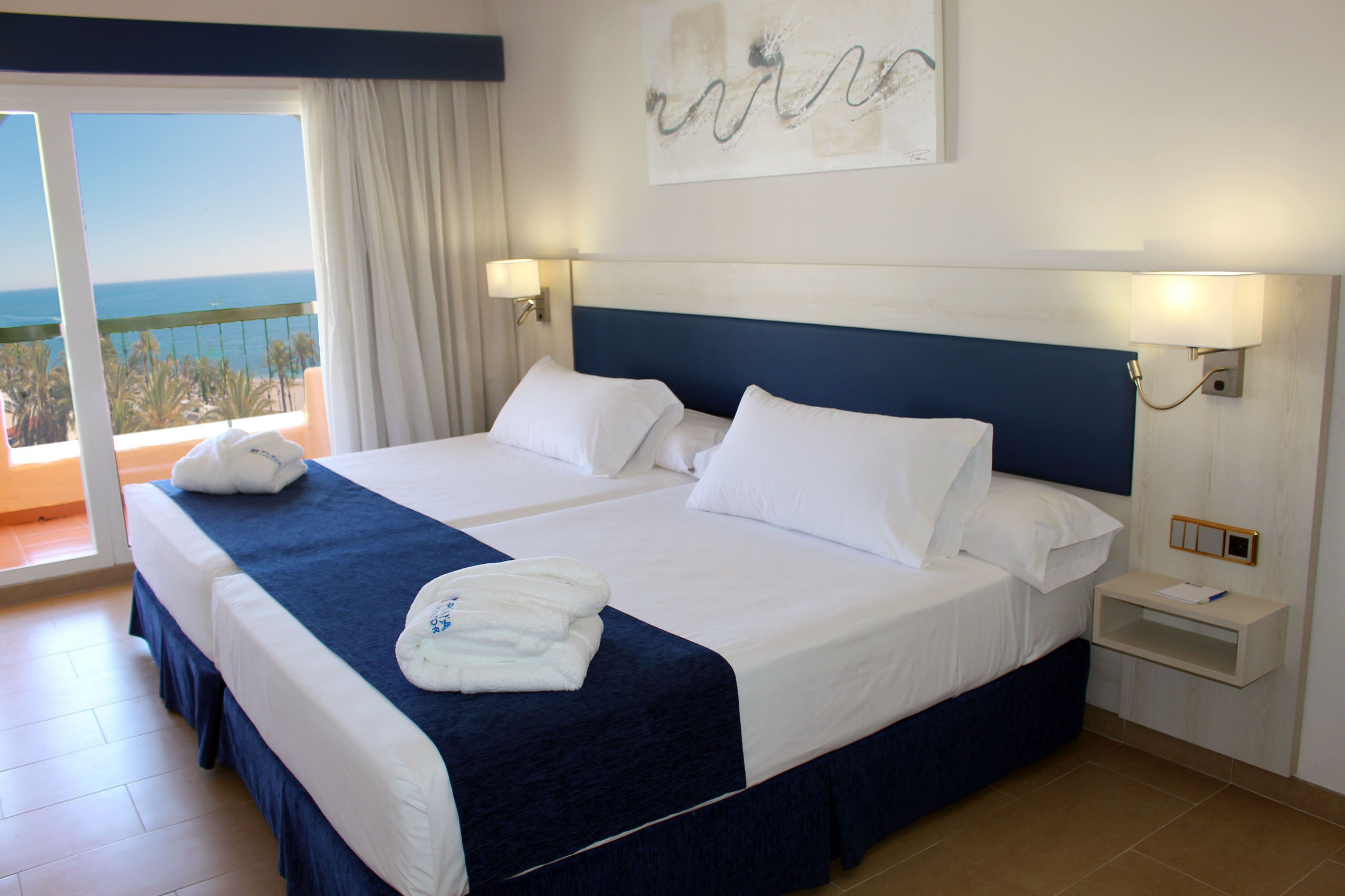 Cama doble con ropa blanca, cabecera azul y vista al océano desde un balcón.