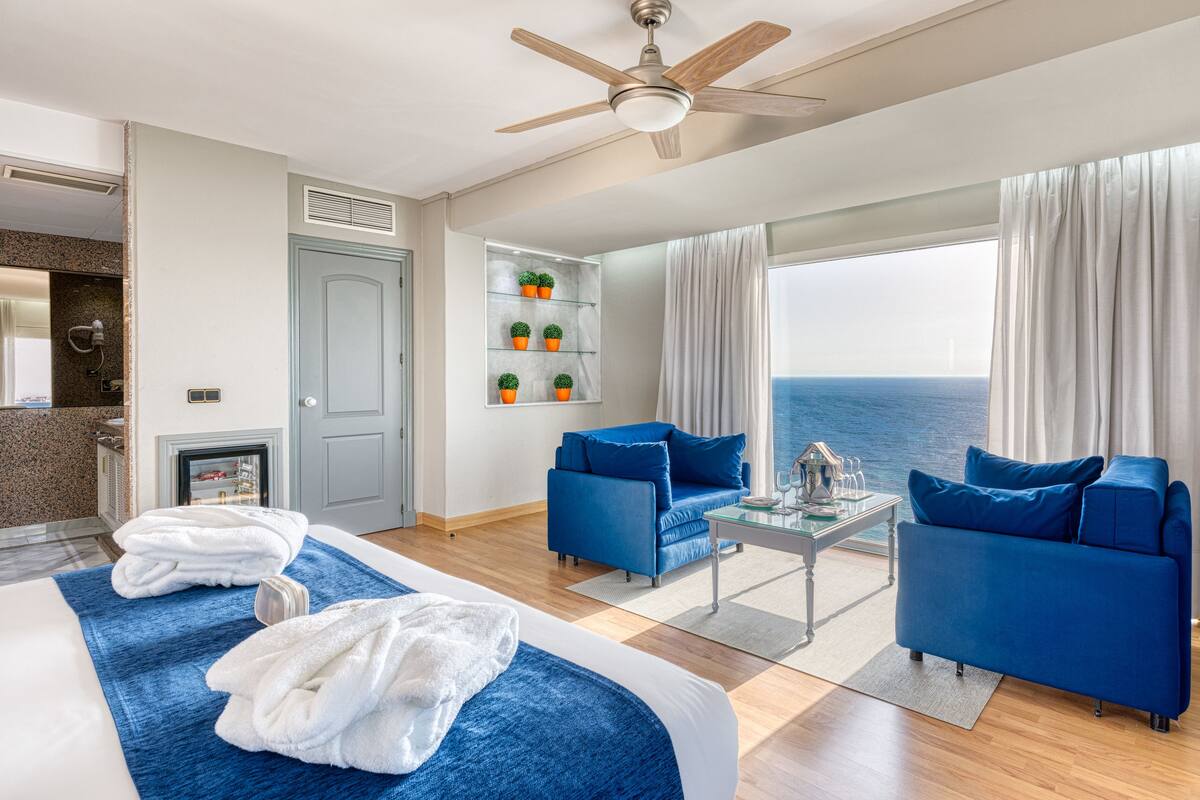 Ein Hotelzimmer mit zwei Betten, einem Deckenventilator, einer Sitzecke mit blauen Sofas, einem kleinen Tisch und Blick aufs Meer.