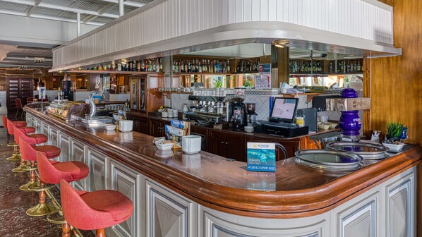 A bar area with a long counter, multiple stools, a coffee machine, and a variety of bottles and glasses.