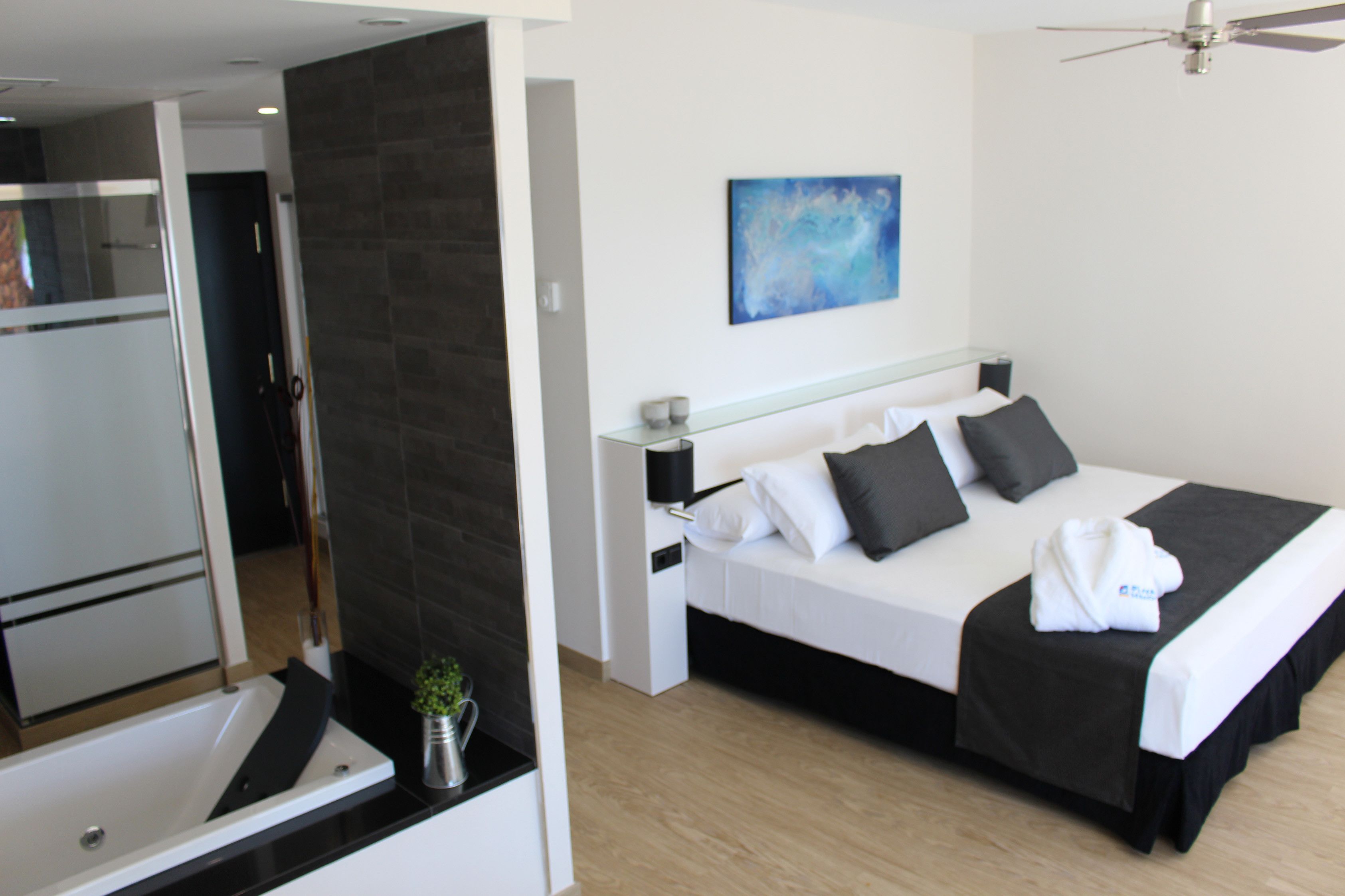 Una habitación de hotel moderna con una cama, una mesita de noche, un ventilador de techo y un baño con bañera y una pequeña planta.