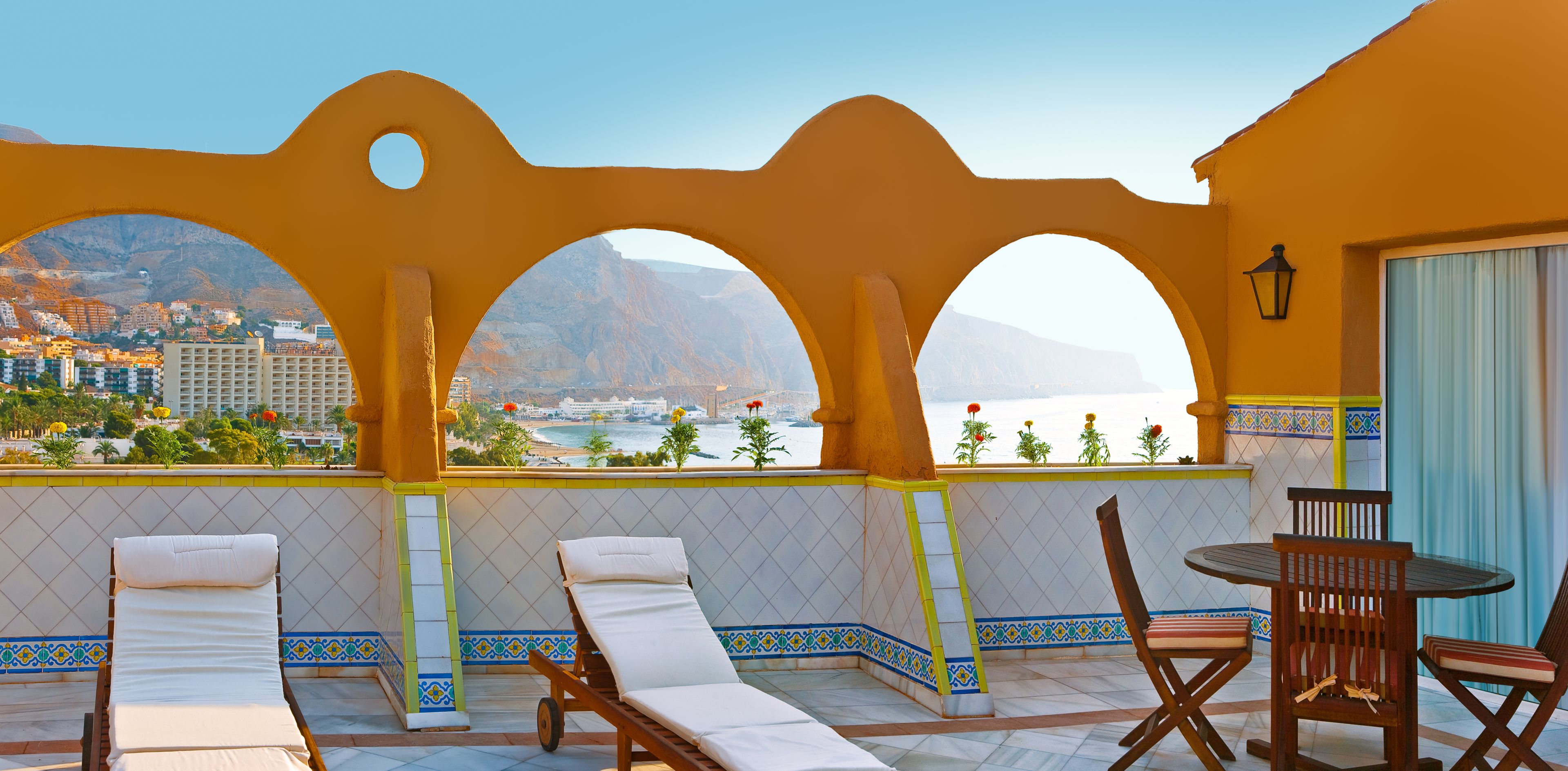 A balcony with a view of a cityscape and mountains, featuring lounge chairs, a table, and a lamp.